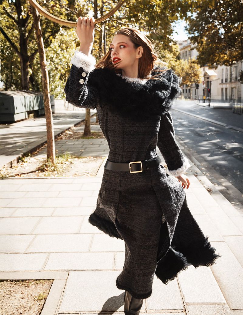 Adult female fashion model in an elegant black coat with fur trim poses on a sidewalk in an urban setting with trees in the background.