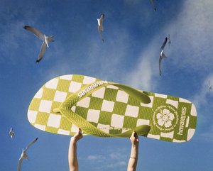 Person holding an oversized Havaianas flip-flop against a blue sky dotted with seagulls in a playful outdoor setting.