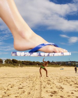 Giant foot hovers over a man on a sandy beach with blue sky, puffy white clouds, ocean view, and other beachgoers in the background.
