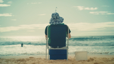 Person sitting on a chair fishing at the beach with ocean waves in the background, wearing a blue and white floral hat.
