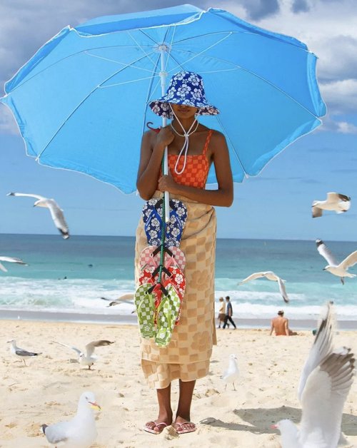 Woman on a sunny beach holding flip-flops, wrapped in a tan towel, under a blue parasol, with seagulls flying over the sand.