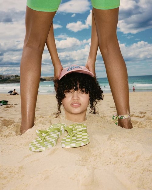 Person buried in sand on a beach with flip-flops and another person standing behind. Ocean and sky with clouds are visible.