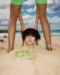 Person with curly hair buried in sand wearing a pink cap next to checkered flip-flops on a beach
