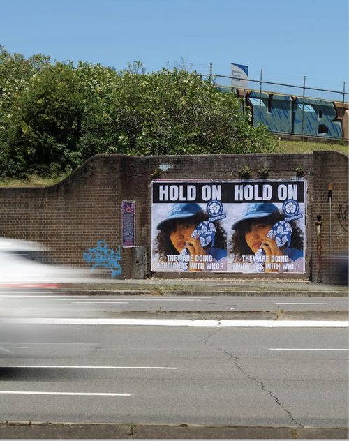 Street view of an outdoor billboard poster on a brick wall with passing car motion blur and green foliage overhead.