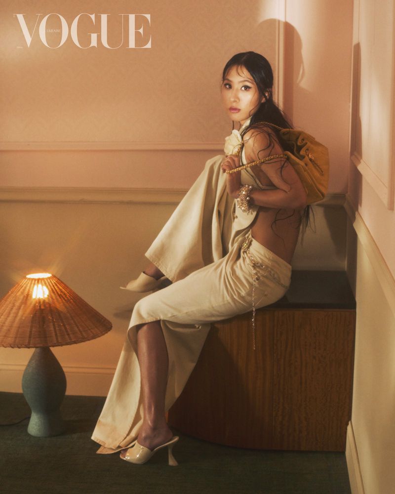 Adult woman poses in a fashion editorial setting while seated, wearing an elegant skirt and a shoulder bag, highlighted by warm lighting.