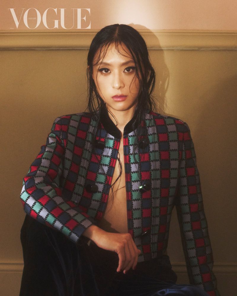 Close-up fashion shot of a person with wet black hair wearing a colourful checkered jacket against a plain tan background.