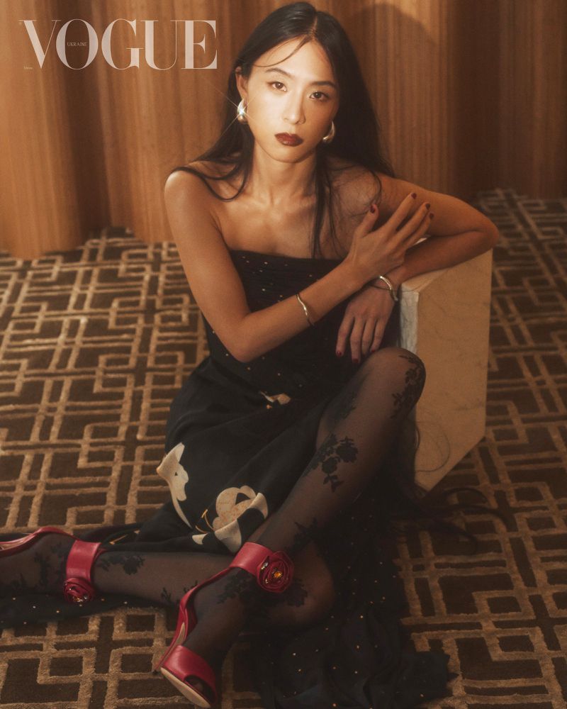 Elegant Asian woman poses in black dress and patterned tights. High heels, jewelry, stylish carpet, neutral wall. Vogue logo visible.