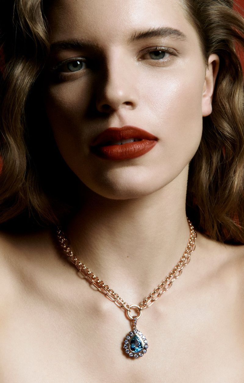 Close-up portrait of a woman with fair skin, wearing dark red lipstick and a gold necklace with a teardrop-shaped blue gemstone pendant.