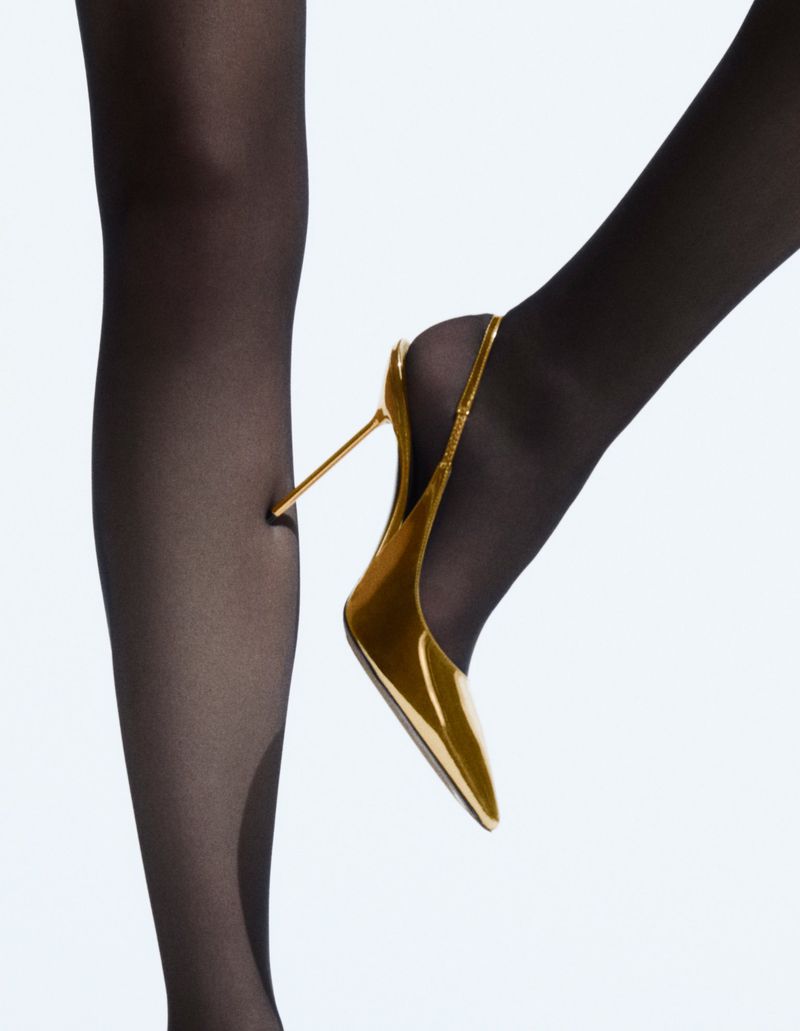 Close-up studio shot of black-stockinged legs, one foot in a gold high-heel pump, against a white background. Fashion shot, leg detail.