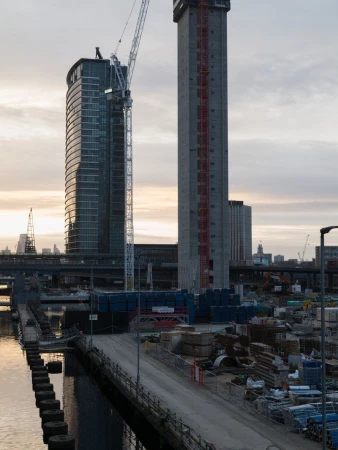 Two modern high-rise buildings stand near a bridge, with a construction crane, waterfront, and sky with light clouds in the background.