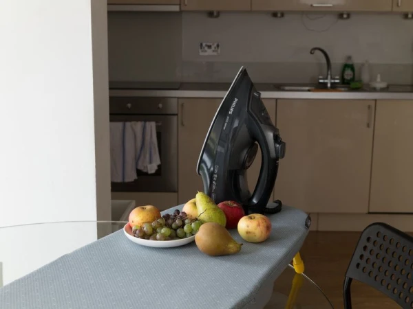 Still life of fruit with an iron, set on an ironing board in a kitchen setting, showcasing a blend of domestic objects and edibles.