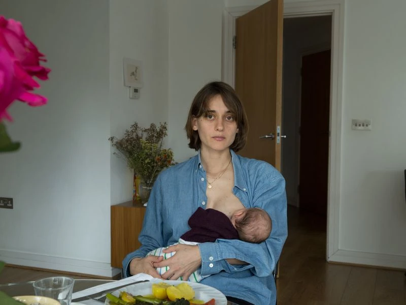 A woman nurses her baby at a table indoors, wearing a blue shirt, with a plate of food and a large pink flower in the foreground.