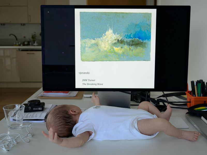 Infant rests on a white desk near a computer monitor displaying a painting, pens, a camera, and a glass of water.