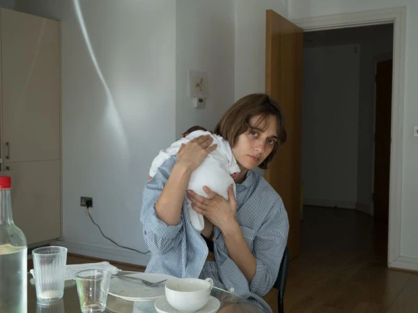 Indoor portrait of a woman holding a baby, seated at a dining table with dishes, bottle and glass, in a brightly lit room.
