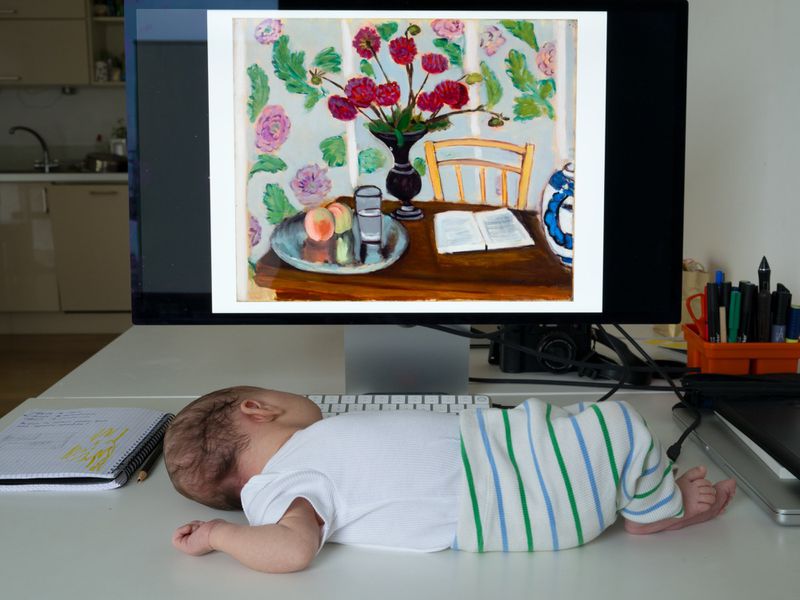 Infant sleeps on white desk near computer monitor displaying colorful still life painting in a well-lit interior room.