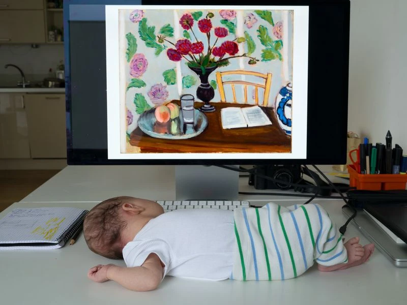 Infant sleeps on white desk near computer monitor displaying colorful still life painting in a well-lit interior room.
