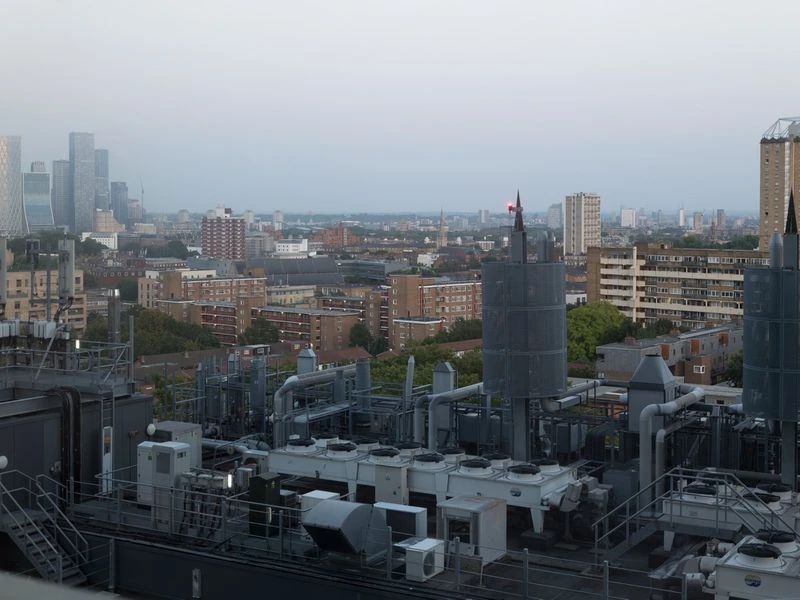 Aerial view of a cityscape featuring dense buildings, industrial rooftop structures, and high-rise skyscrapers in the hazy distance.
