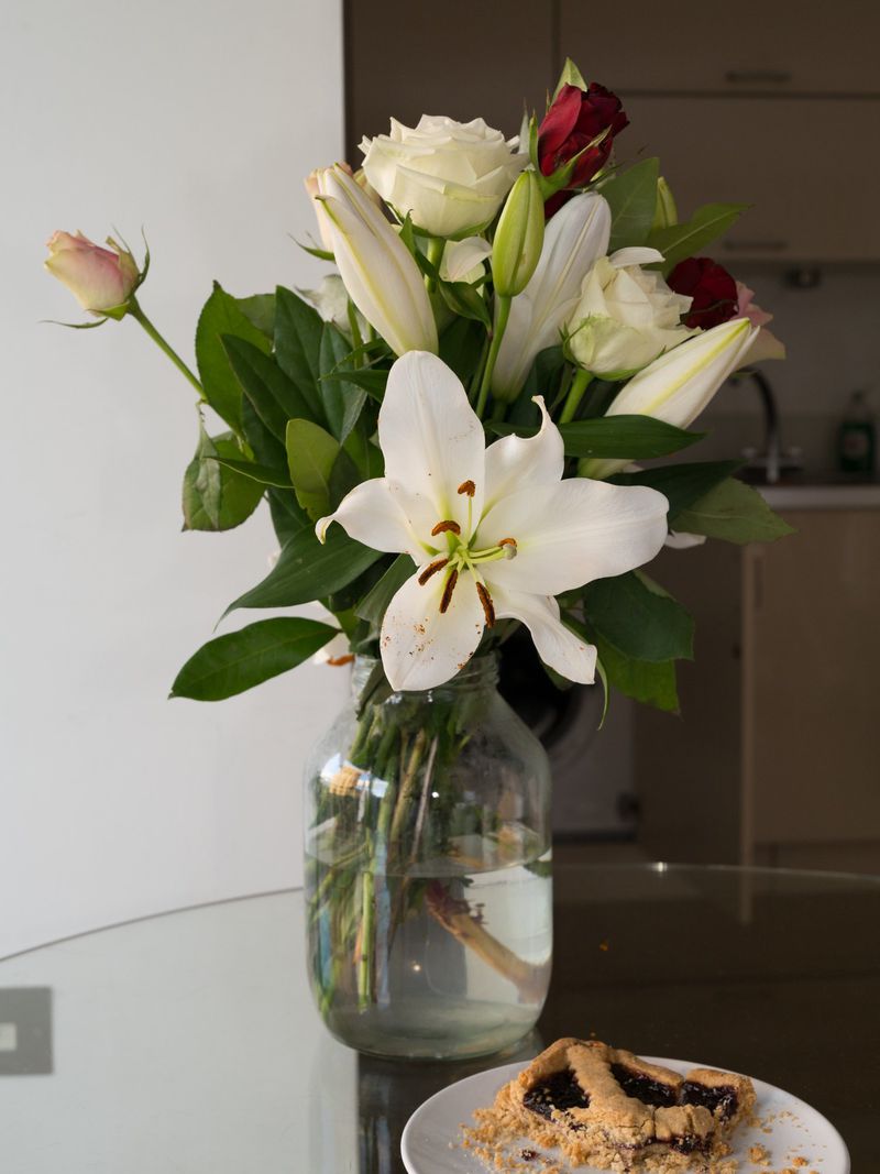 Bouquet of roses and lilies in a glass jar on a glass table next to a pastry, set in a home interior environment.