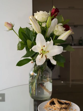 Bouquet of roses and lilies in a glass jar on a glass table next to a pastry, set in a home interior environment.