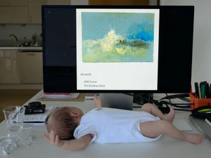 A baby wearing a white bodysuit lies on a white desk in front of a computer monitor displaying artwork.