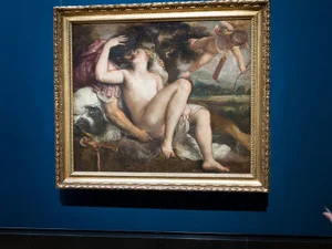 Classical oil painting with embracing figures in an ornate gilded gold frame hanging against a deep blue gallery wall.