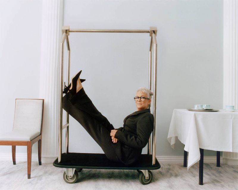 Mature woman with short grey hair, wearing glasses and a dark suit, lies back in a luggage cart in an indoor environment.