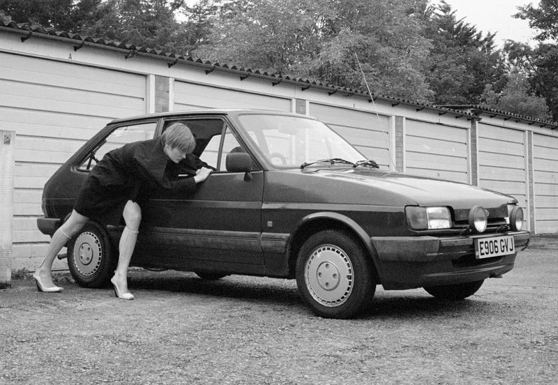 Black-and-white shot of a woman in heels pushing a black car near a building and trees, creating a stark contrast and vintage feel.