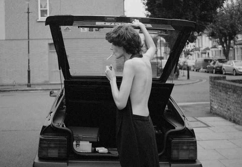 Black and white medium shot of a slender woman with dark hair standing near a car with its trunk open, smoking a cigarette on a city street.