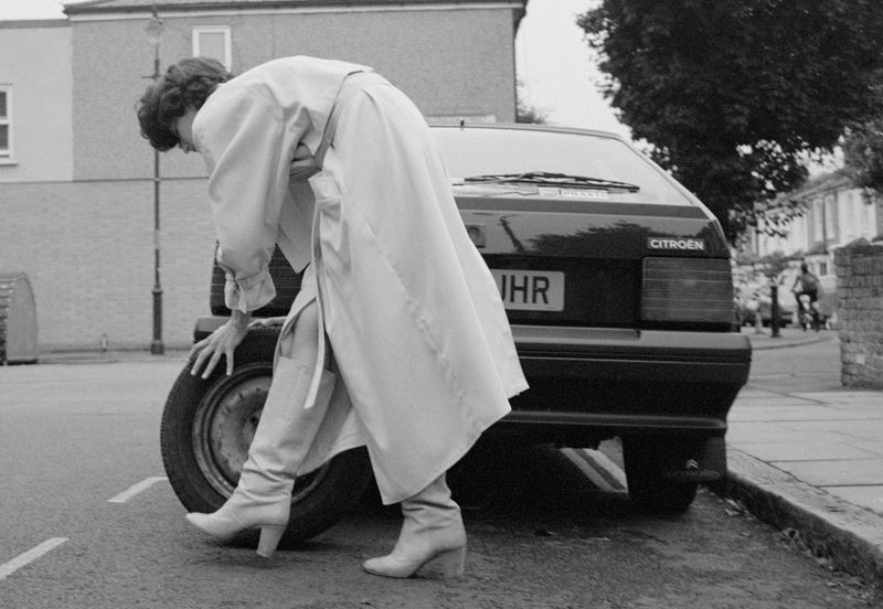 Black and white shot of a woman changing the tire on a black Citroen car on a city street. Boots and a long coat are visible.