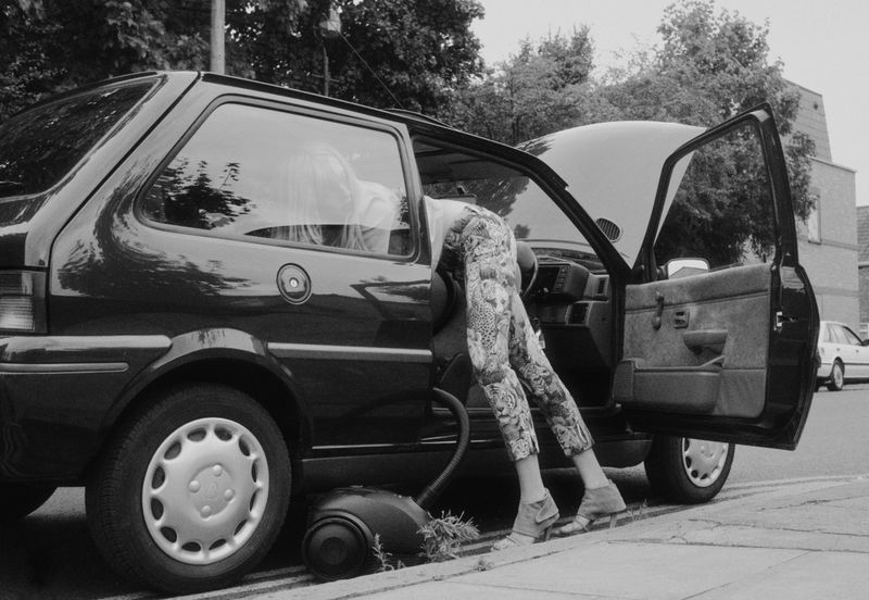 Black and white image of a woman in patterned pants with her upper body inside the hood of a car and a vacuum cleaner nearby.