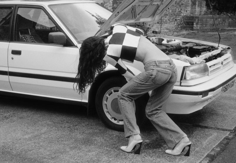 Woman in jeans and checkered top looks under the hood of a white car on a paved lot with a partial brick wall in the background.