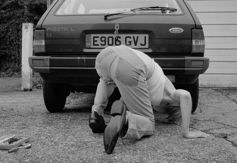An adult female in denim overalls is bent over, looking at a Ford Fiesta on an outdoor concrete surface with tools nearby.