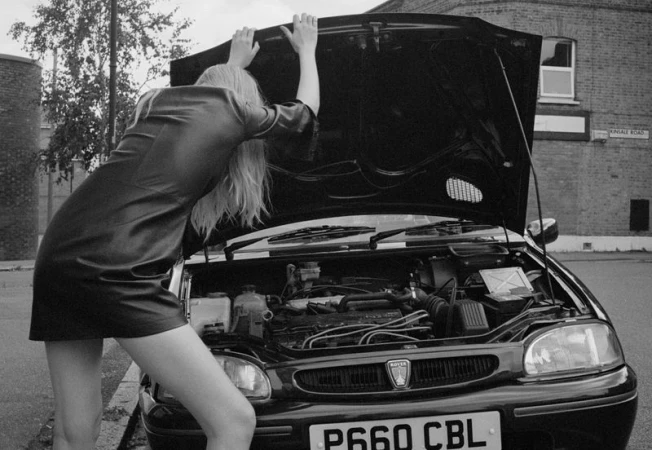 A black and white photograph of a woman with long blonde hair looking under the open hood of a black Rover car on a street.