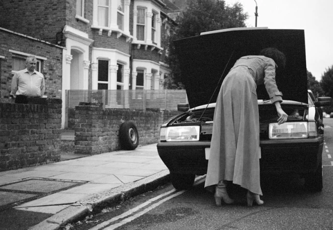 Woman in long coat leaning into car engine compartment; adult man standing near brick wall; tire visible in the background.