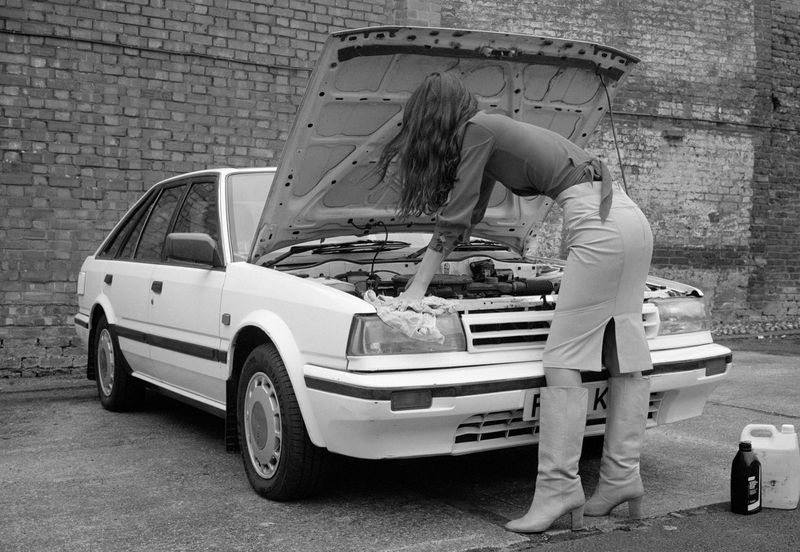 A woman is working on the engine of a white car in front of a brick wall with oil and cleaning cloth, showcasing an automotive scene.