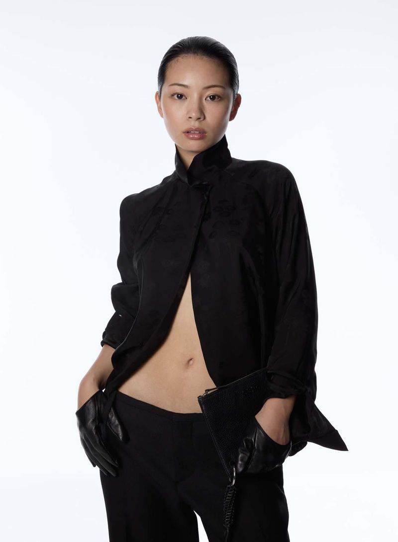 Asian woman in black jacket and pants poses in a photo studio wearing leather gloves and holding a black clutch, illuminated by studio lighting.