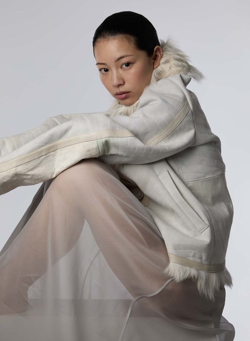 An Asian woman with a shaved head poses indoors, wearing a fur-trimmed jacket and sheer skirt, in a fashion-forward studio portrait.