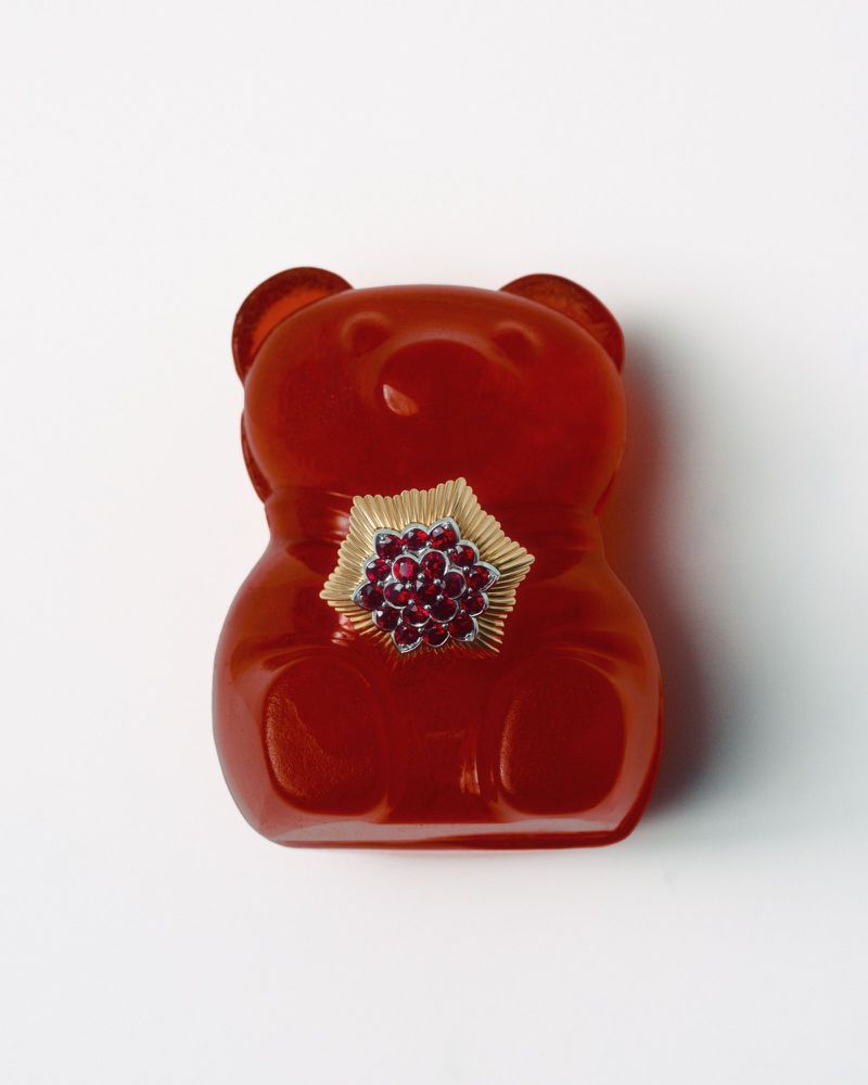 Red gummy bear sculpture with a flower brooch. The gummy bear is smooth and translucent. The brooch has gold leaves and red gems.