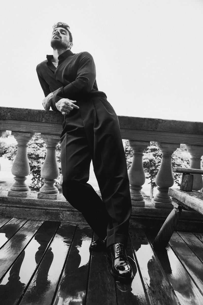 Monochrome shot of a man in a black turtleneck and pants leaning against a stone railing, with a wet, reflective patio floor.