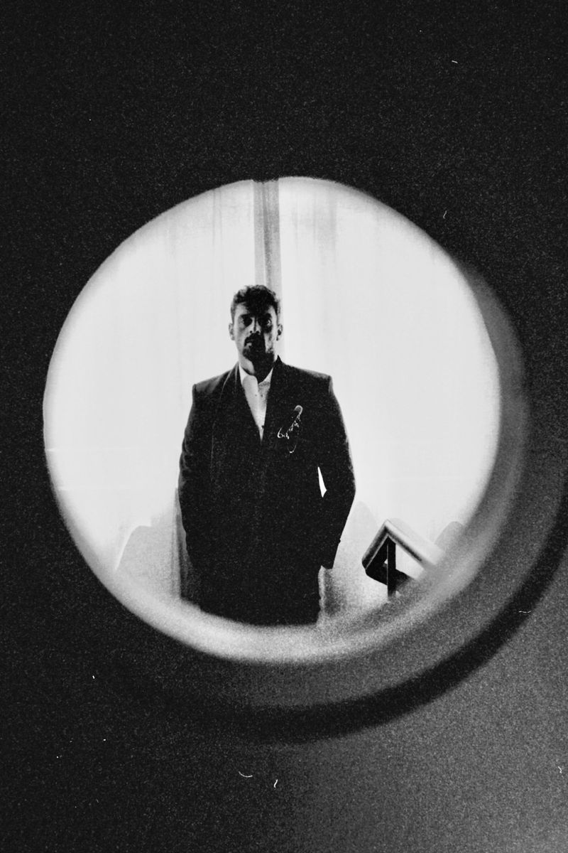 Black and white portrait of an adult man in a dark suit, framed by a circular window, looking directly at the camera.