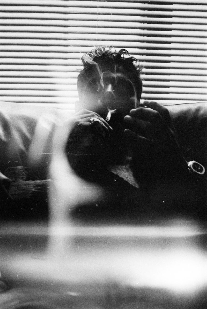Black-and-white portrait of an adult male with a cigarette indoors, showcasing smoke and Venetian blinds.