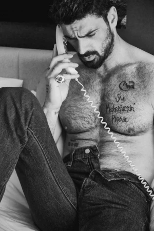 Black and white portrait of a man with beard, talking on a vintage telephone in an indoor setting, wearing jeans.