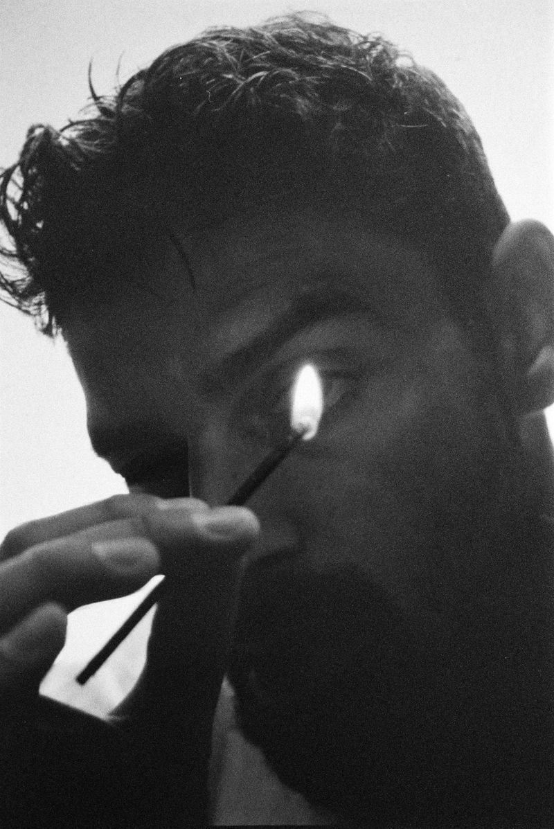 Monochrome portrait of an adult male with a lit match near his eye. Dark hair, and grayscale rendering create a dramatic close-up composition.