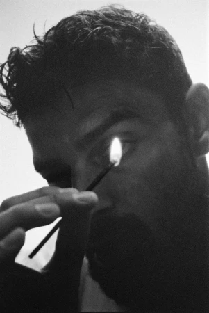 Monochrome portrait of an adult male with a lit match near his eye. Dark hair, and grayscale rendering create a dramatic close-up composition.