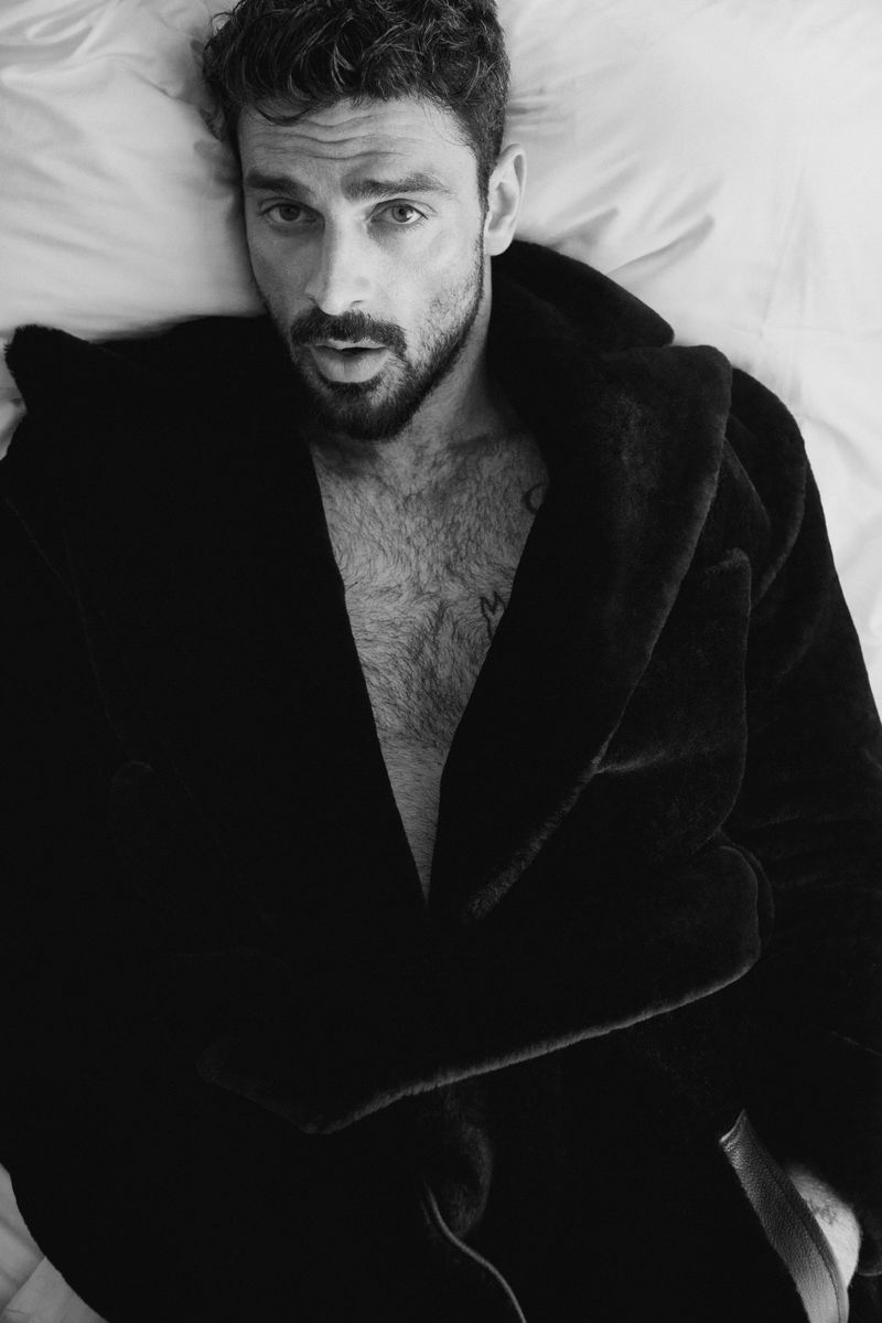 Black and white portrait of a man with dark curly hair and a beard, lying on white bedding wearing a dark robe.
