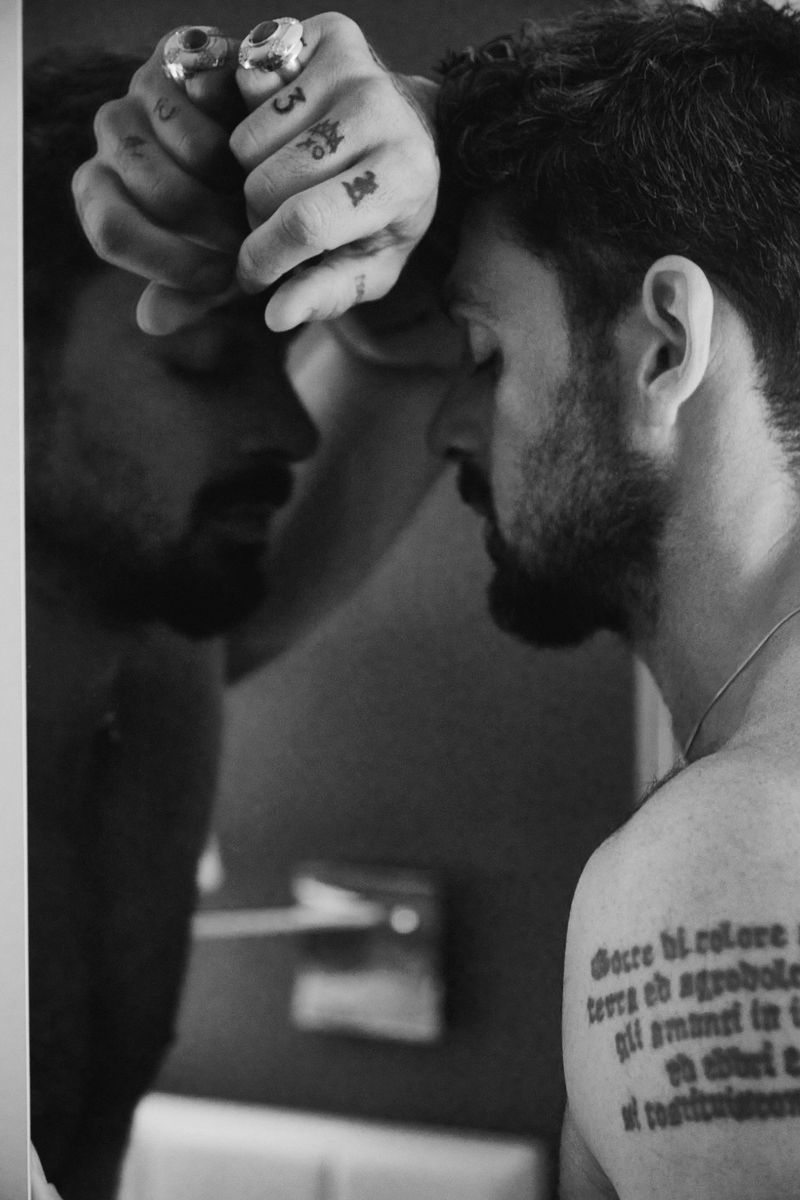 Monochromatic close-up of a man with a beard leaning his head on a mirror, his face partially visible with hand tattoos and rings.