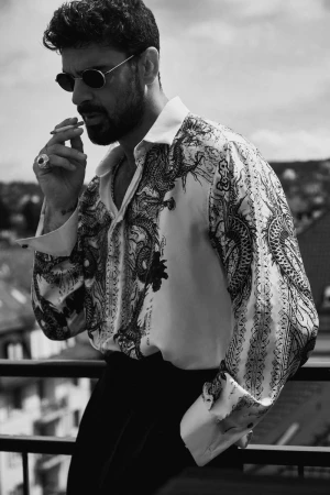 Black and white portrait of Michele Morrone in a printed silk shirt and sunglasses holding a cigarette on a balcony.