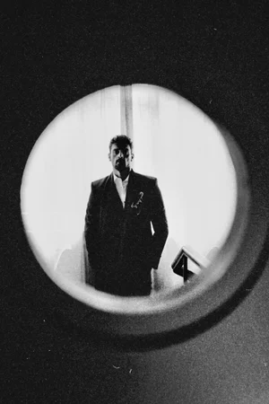 Monochrome portrait of actor Michele Morrone wearing a dark tailored suit, viewed through a circular peephole cutout.