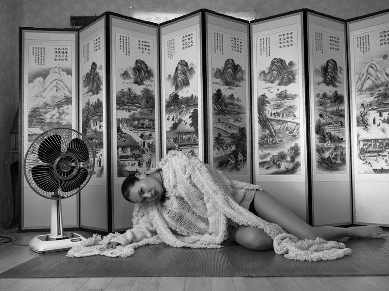 Black and white portrait of a woman reclining in a textured robe next to a retro fan in front of an Asian-style folding screen.