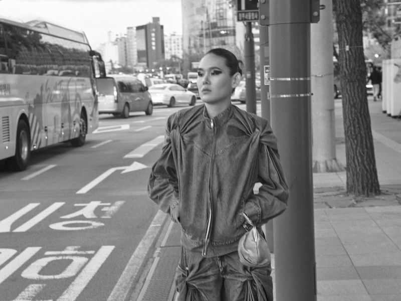 Model EZ stands on a city sidewalk wearing a structured denim jacket with gathered shoulder details beside a street pole.